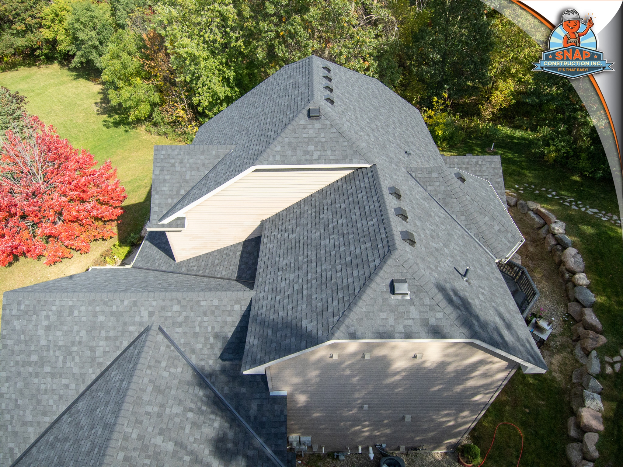 Aerial view of asphalt shingle home roof, roof repair by Snap Construction in Bloomington MN, showing vents and ridges amid fall trees
