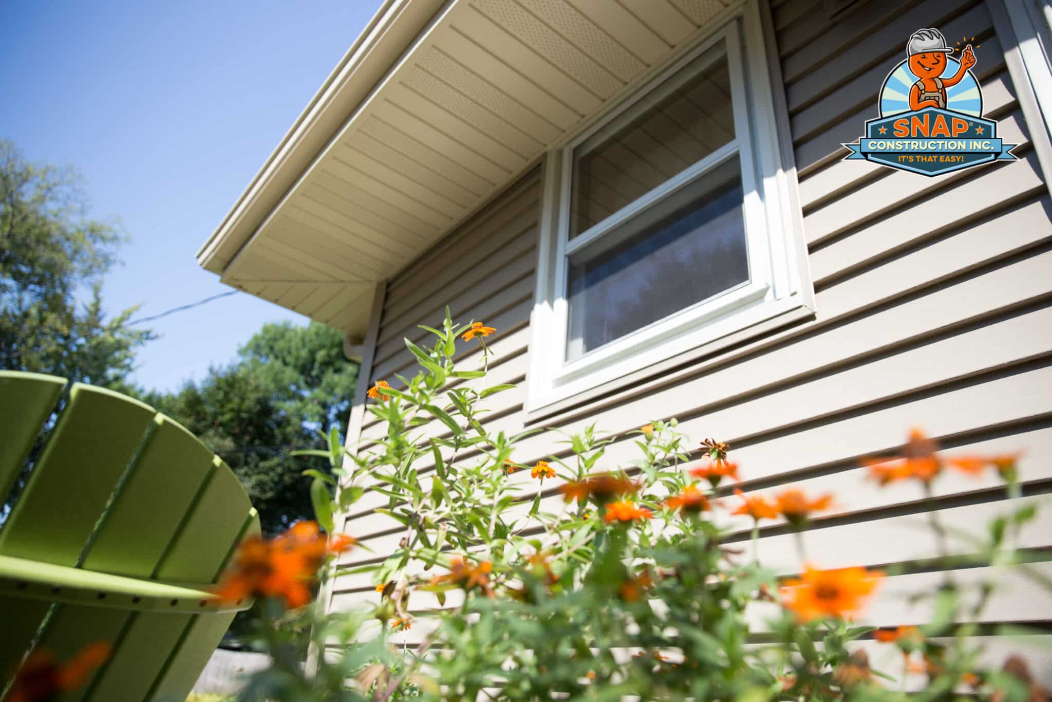 Snap Construction siding contractor in Bloomington MN showcasing tan vinyl siding, soffit and window with flowers under blue sky