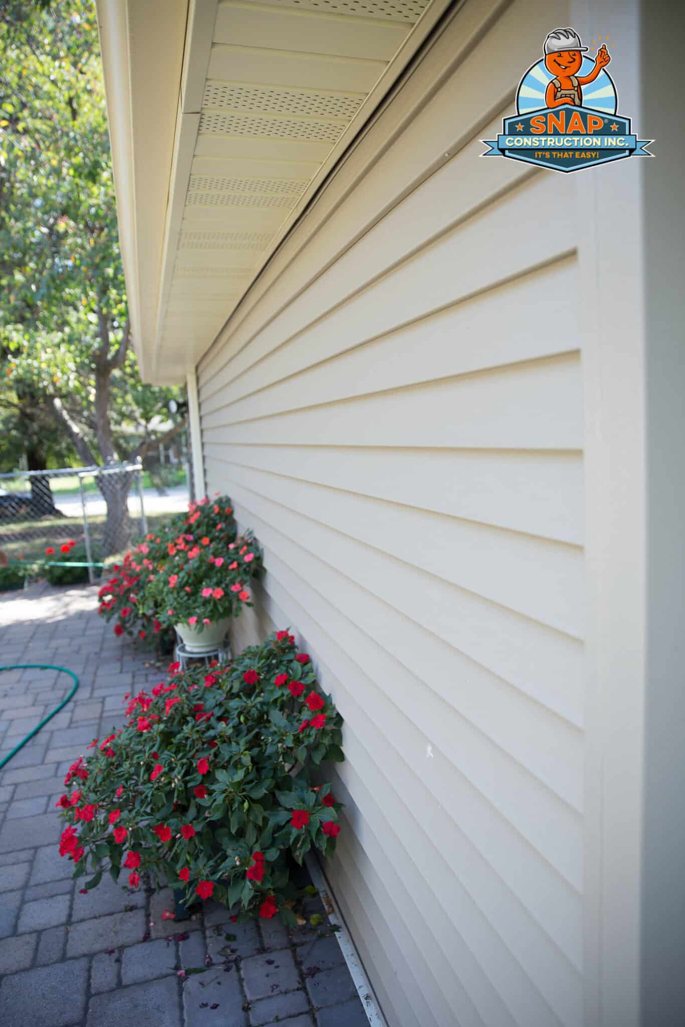 Fresh beige vinyl siding replacement by Snap Construction on a Bloomington MN home, with soffits, paver patio, and red flowers along wall