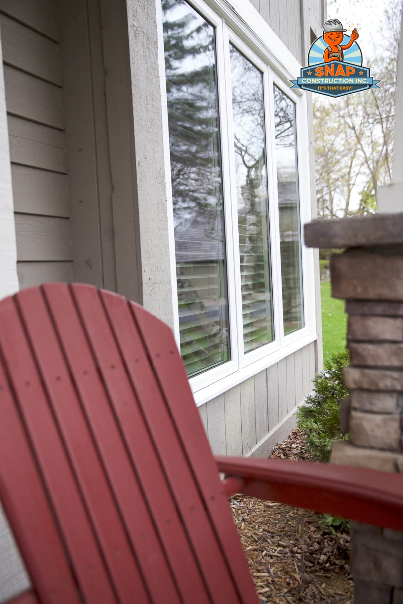 Snap Construction vinyl windows on gray siding in Bloomington MN, viewed from patio with red Adirondack chair and stone column